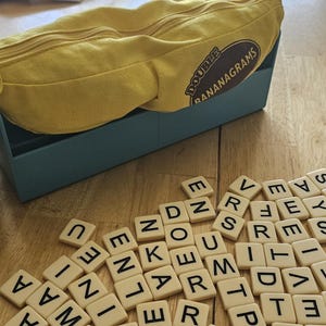 May include: A yellow Bananagrams game set with a banana-shaped pouch and a blue container. Scattered letter tiles are on a wooden surface. The pouch has the text "DOUBLE BANANAGRAMS".