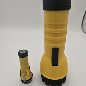 Może przedstawiać: Dwie żółte latarki z czarnymi akcentami. Większa latarka ma teksturowany uchwyt i czarny pasek. Mniejsza latarka ma pierścień przymocowany do góry.