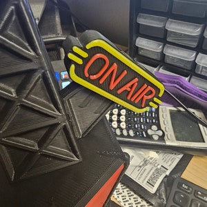 Puede incluir: Un letrero "ON AIR" negro y amarillo con letras rojas. La imagen tambi&eacute;n muestra un objeto geom&eacute;trico negro con detalles rojos, una calculadora y un teclado. Ideal para estudios.