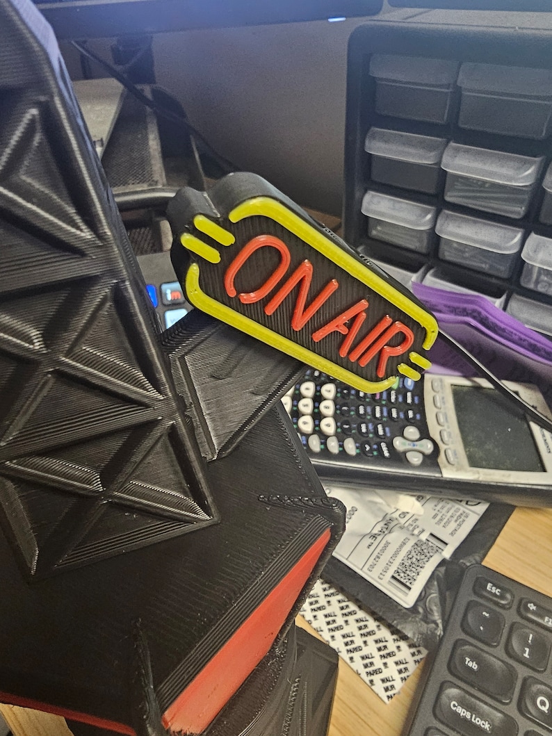 Puede incluir: Un letrero "ON AIR" negro y amarillo con letras rojas. La imagen tambi&eacute;n muestra un objeto geom&eacute;trico negro con detalles rojos, una calculadora y un teclado. Ideal para estudios.
