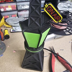 Puede incluir: Una l&aacute;mpara negra y verde impresa en 3D con un letrero "ON AIR". La l&aacute;mpara tiene una estructura similar a una torre con un centro verde iluminado. El letrero es amarillo con texto rojo. La l&aacute;mpara est&aacute; sobre una superficie de trabajo.