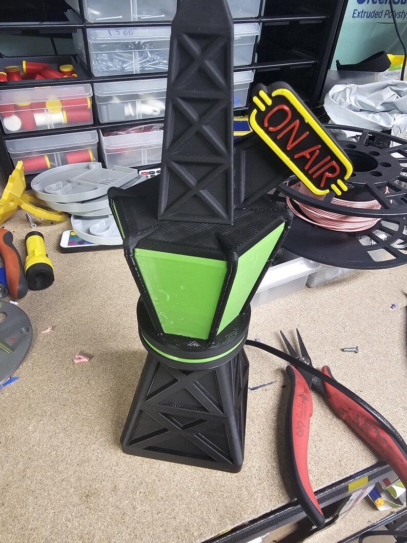 Puede incluir: Una l&aacute;mpara negra y verde impresa en 3D con un letrero "ON AIR". La l&aacute;mpara tiene una estructura similar a una torre con un centro verde iluminado. El letrero es amarillo con texto rojo. La l&aacute;mpara est&aacute; sobre una superficie de trabajo.
