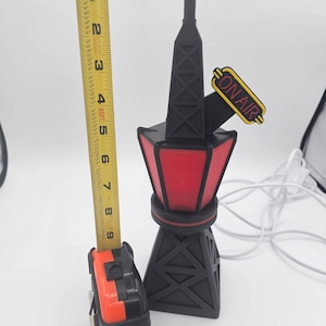 Puede incluir: Una l&aacute;mpara con forma de torre de radio negra y roja con las palabras "ON AIR" en amarillo. La l&aacute;mpara mide aproximadamente 23 cm de altura y tiene una base y parte superior negras con un centro rojo iluminado.