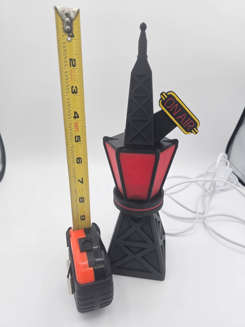 Puede incluir: Una l&aacute;mpara con forma de torre de radio negra y roja con las palabras "ON AIR" en amarillo. La l&aacute;mpara mide aproximadamente 23 cm de altura y tiene una base y parte superior negras con un centro rojo iluminado.