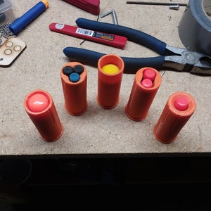 May include: Five orange plastic tubes with different colored round objects inside. The tubes are arranged in a row on a wooden surface.
