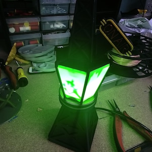 Puede incluir: Una l&aacute;mpara negra y verde iluminada con un dise&ntilde;o geom&eacute;trico. La l&aacute;mpara tiene una base cuadrada y una parte superior de m&uacute;ltiples lados con una luz verde brillante. Las palabras "ON AIR" son visibles en un letrero amarillo. Alicates y otras herramientas est&aacute;n en el fondo.