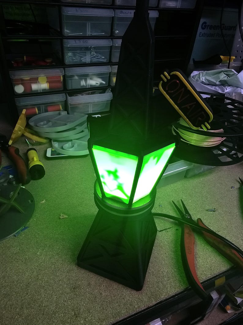 Puede incluir: Una l&aacute;mpara negra y verde iluminada con un dise&ntilde;o geom&eacute;trico. La l&aacute;mpara tiene una base cuadrada y una parte superior de m&uacute;ltiples lados con una luz verde brillante. Las palabras "ON AIR" son visibles en un letrero amarillo. Alicates y otras herramientas est&aacute;n en el fondo.