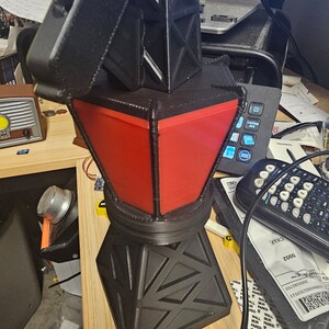 Puede incluir: Una l&aacute;mpara geom&eacute;trica negra y roja con un dise&ntilde;o &uacute;nico. La l&aacute;mpara tiene una base en forma de diamante y una secci&oacute;n central roja trapezoidal. La parte superior es negra y angular. La l&aacute;mpara est&aacute; enchufada a una toma de corriente.