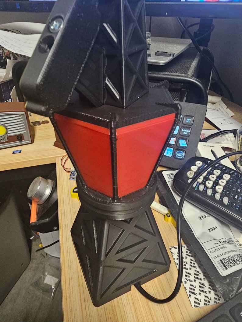 Puede incluir: Una l&aacute;mpara geom&eacute;trica negra y roja con un dise&ntilde;o &uacute;nico. La l&aacute;mpara tiene una base en forma de diamante y una secci&oacute;n central roja trapezoidal. La parte superior es negra y angular. La l&aacute;mpara est&aacute; enchufada a una toma de corriente.