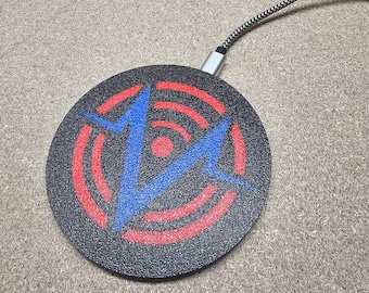 Puede incluir: Una base de carga inalámbrica negra con un diseño de rayo rojo y azul. La base de carga tiene un cable trenzado negro.