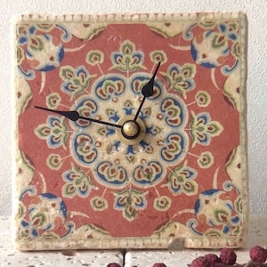 Könnte beinhalten: Quadratische Uhr mit einem roten, blauen und grünen Blumenmuster. Das Zifferblatt hat schwarze Zeiger und eine goldfarbene Mitte. Die Uhr hat einen cremefarbenen Rand.