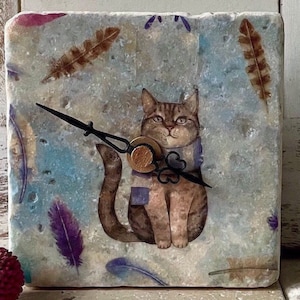 Könnte beinhalten: Eine quadratische, hellblaue und weiße Uhr mit einer braunen Katze, die einen blauen Schal trägt. Die Uhr hat schwarze Zeiger und einen Holzknopf. Der Hintergrund ist mit Federn verziert.