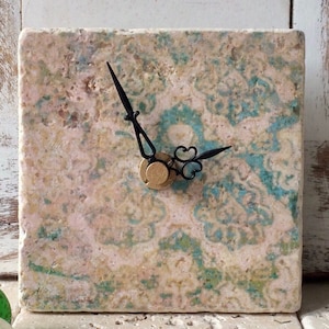 Könnte beinhalten: Eine quadratische Uhr mit schwarzen Zeigern auf einem beigefarbenen Hintergrund mit grünen und blauen Marmorierungen. Die Uhr ist aus Stein.