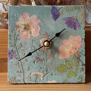Könnte beinhalten: Eine quadratische Wanduhr mit einem hellblauen, strukturierten Zifferblatt, das mit eingearbeiteten gepressten Blumen in Rosa- und Violetttönen verziert ist. Die Uhr hat zwei dünne schwarze Zeiger an einem kleinen goldenen Mechanismus.