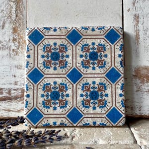 Könnte beinhalten: Quadratischer Keramikuntersetzer mit einem sich wiederholenden Muster aus blauen und braunen floralen Designs auf cremefarbenem Hintergrund. Der Untersetzer hat ein blaues Rautenmuster an den Rändern.