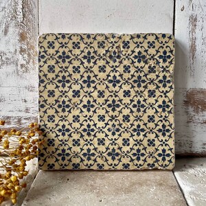 Könnte beinhalten: Eine quadratische Fliese mit einem sich wiederholenden Muster aus blauen floralen Designs auf beigefarbenem Hintergrund.