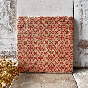Könnte beinhalten: Quadratischer Untersetzer mit beigem Hintergrund und sich wiederholendem rotem Blumenmuster. Der Untersetzer hat eine strukturierte Oberfläche und besteht aus einem steinartigen Material. Das Design weist eine klassische, Vintage-Ästhetik auf.