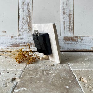 May include: A small, off-white rectangular clock face with a black clock mechanism attached to the back. The clock is propped up on a small, light-coloured wooden stand. Dried yellow flowers are in the foreground.