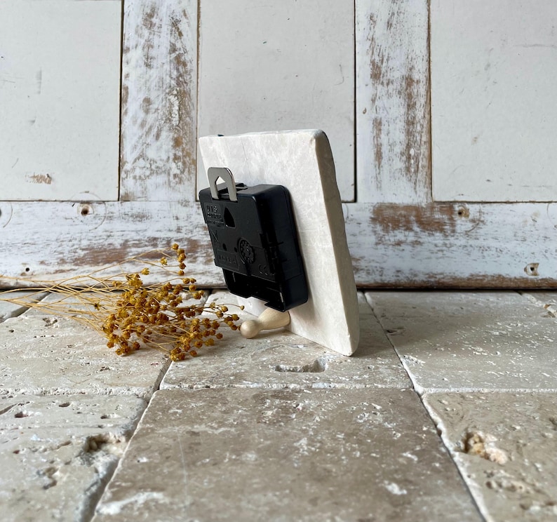May include: A small, off-white rectangular clock face with a black clock mechanism attached to the back. The clock is propped up on a small, light-coloured wooden stand. Dried yellow flowers are in the foreground.