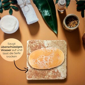 May include: An oval orange soap bar with white lather rests on a square, porous, beige stone soap dish. A circular text bubble points to the stone, featuring the German text: Saugt &uuml;bersch&uuml;ssiges Wasser auf und l&auml;sst die Seife trocken.