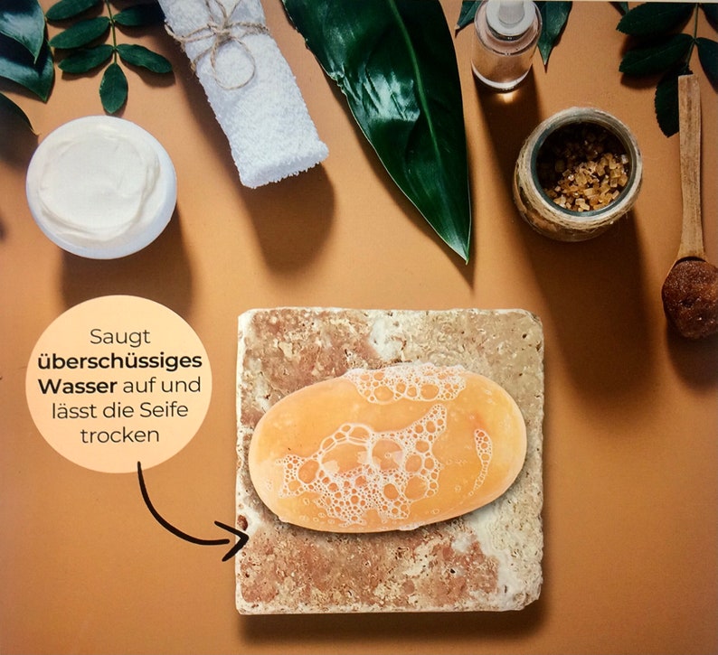 May include: An oval orange soap bar with white lather rests on a square, porous, beige stone soap dish. A circular text bubble points to the stone, featuring the German text: Saugt &uuml;bersch&uuml;ssiges Wasser auf und l&auml;sst die Seife trocken.