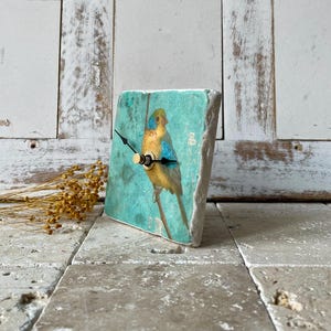 May include: A square decorative clock featuring a bird illustration. The clock face is a light teal colour, displaying a yellow and blue bird. The clock hands are black with a gold-coloured centre. The clock is made from a stone-like material.