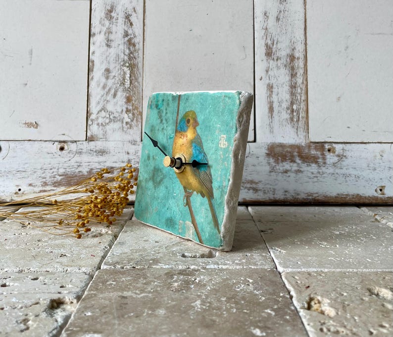 May include: A square decorative clock featuring a bird illustration. The clock face is a light teal colour, displaying a yellow and blue bird. The clock hands are black with a gold-coloured centre. The clock is made from a stone-like material.