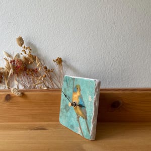May include: A square clock featuring a painted bird on a branch against a turquoise backdrop. The clock has black hands and a wooden frame. It is displayed on a wooden surface. The clock's dimensions are approximately 15 cm x 15 cm.