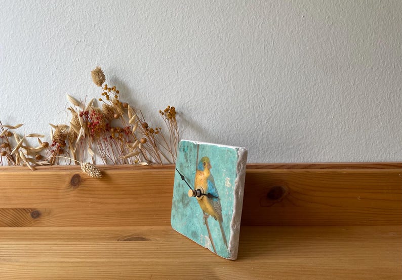 May include: A square clock featuring a painted bird on a branch against a turquoise backdrop. The clock has black hands and a wooden frame. It is displayed on a wooden surface. The clock's dimensions are approximately 15 cm x 15 cm.