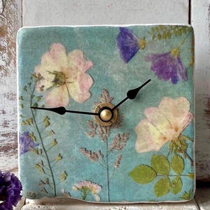 Könnte beinhalten: Quadratische Uhr mit floralem Design. Das Zifferblatt ist hellblau mit gepressten Blumen in Rosa, Lila und Grün. Die Zeiger sind schwarz und die Mitte ist goldfarben.