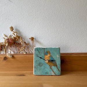 May include: Square ceramic clock with a blue and yellow bird illustration. The clock has black hands and a wooden base. The background is a light teal colour. Dried flowers are arranged to the left of the clock.