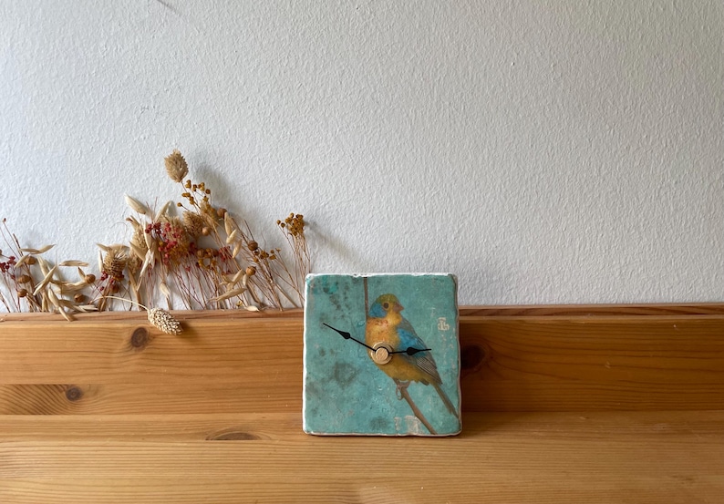 May include: Square ceramic clock with a blue and yellow bird illustration. The clock has black hands and a wooden base. The background is a light teal colour. Dried flowers are arranged to the left of the clock.