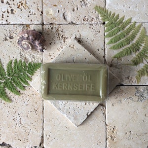 May include: A rectangular olive green bar of soap wrapped in clear plastic, embossed with the text OLIVEN&Ouml;L KERNSEIFE. The soap is positioned on a square stone tile, accompanied by two green fern leaves and a small, textured brown seashell.