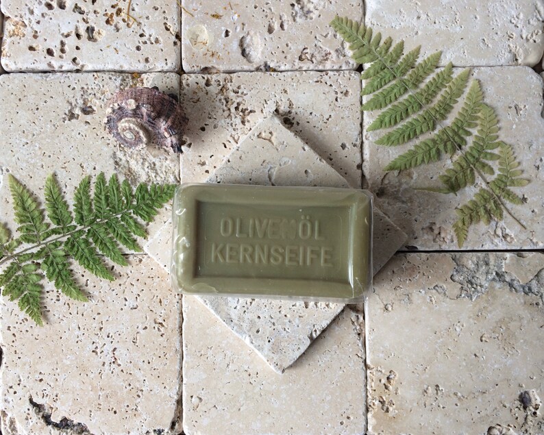 May include: A rectangular olive green bar of soap wrapped in clear plastic, embossed with the text OLIVEN&Ouml;L KERNSEIFE. The soap is positioned on a square stone tile, accompanied by two green fern leaves and a small, textured brown seashell.