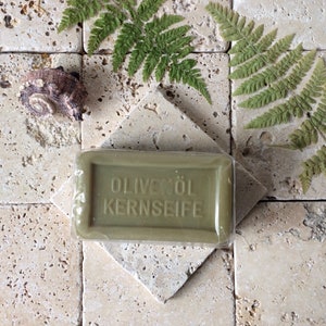 May include: A rectangular bar of olive oil soap wrapped in clear plastic film. The soap is olive green and features the embossed text OLIVEN&Ouml;L KERNSEIFE on its front surface. The bar is positioned on a square, light-colored, textured stone tile.