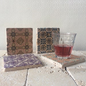 Könnte beinhalten: Vier quadratische Untersetzer aus Keramik mit verschiedenen Mustern. Die Untersetzer befinden sich auf einer hellbeigen Oberfläche. Ein Glas mit rosa Flüssigkeit steht auf einem der Untersetzer.