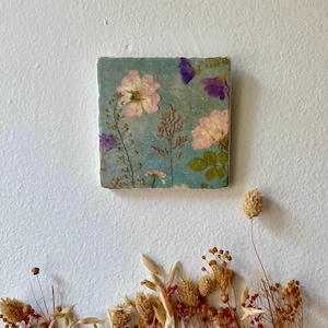 Könnte beinhalten: Eine quadratische Fliese mit einem gepressten Blumenmuster. Die Fliese zeigt rosa und violette Blumen auf blauem Hintergrund.
