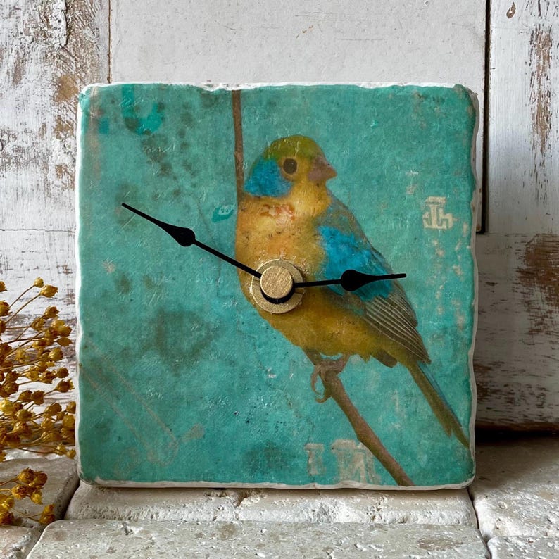 May include: Square decorative clock with a bird illustration. The bird has yellow, blue, and green plumage, perched on a branch against a teal background. The clock has black hands and a gold-coloured centre.