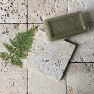 May include: A rectangular bar of olive oil soap in clear plastic packaging with the embossed text OLIVEN&Ouml;L KERNSEIFE, accompanied by a single green fern leaf. The soap is a muted olive green color with a smooth, solid, and firm appearance.