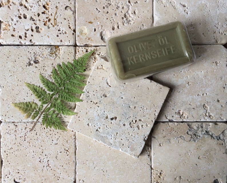 May include: A rectangular bar of olive oil soap in clear plastic packaging with the embossed text OLIVEN&Ouml;L KERNSEIFE, accompanied by a single green fern leaf. The soap is a muted olive green color with a smooth, solid, and firm appearance.