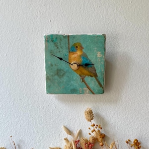 May include: Square wall clock with a bird illustration on a teal background. The clock has black hands and a gold-coloured centre. The clock is mounted on a white wall, with dried flowers at the bottom.