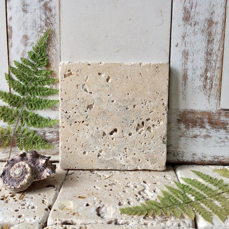 May include: A square, beige travertine tile with a porous, pitted surface texture. The natural stone features irregular holes and crevices throughout its flat face, showcasing the organic, earthy appearance of the material in a neutral tone.