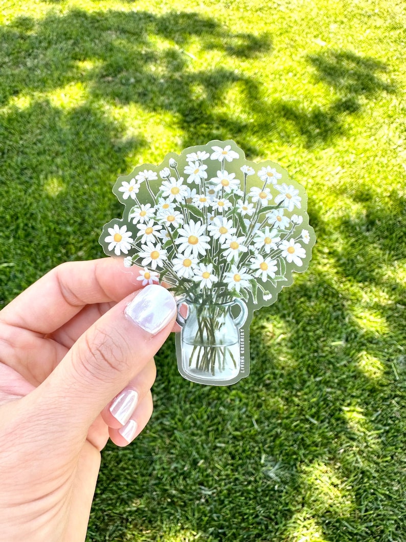 May include: A clear glass vase filled with a bouquet of white daisies. The vase has two handles and the text "Living Greenly" is printed on the side.