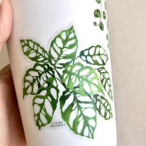 May include: A white sticker featuring a green illustration of a plant with large, patterned leaves. The sticker reads "LIVING GREENERY" at the bottom.