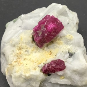 Op de afbeelding: Twee rode robijnkristallen ingebed in een witte rots. De robijnkristallen zijn scherp en hebben een dieprode kleur.