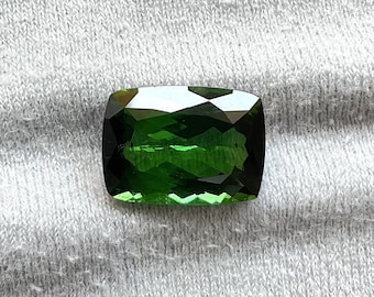 May include: A deep green gemstone with a rectangular cut and faceted edges. The stone is set against a white fabric background.