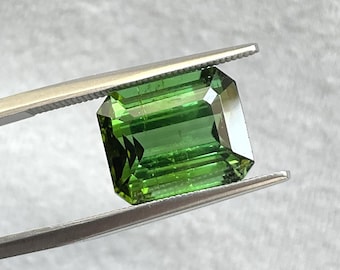 May include: A vibrant green gemstone, likely a tourmaline, is held by tweezers. The gemstone is cut into a rectangular emerald shape with facets.