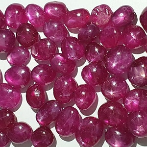 May include: A collection of pink star ruby gemstones, each with a unique star pattern. The stones are smooth and polished, with a slightly rounded shape.