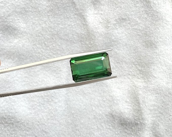 May include: A vibrant green emerald gemstone, cut in an emerald shape, is held by a pair of tweezers against a white background.