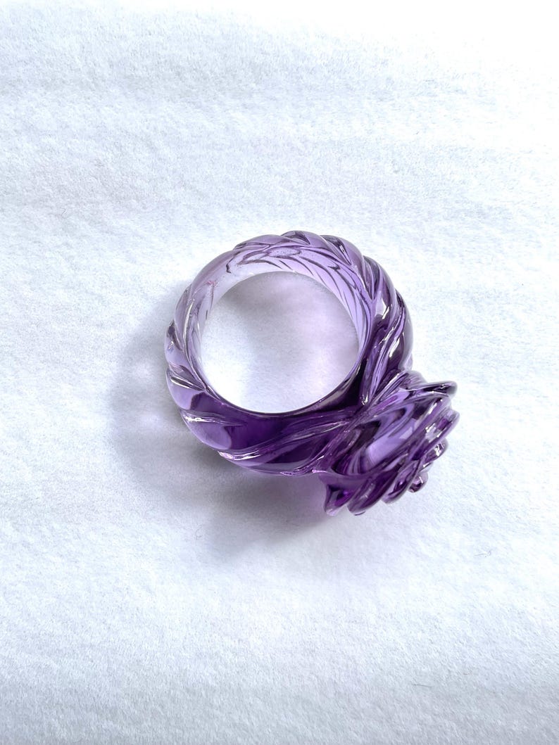 Pode incluir: Um anel de ametista roxo com um design de rosa. O anel &eacute; esculpido em uma &uacute;nica pe&ccedil;a de ametista e tem uma banda torcida.